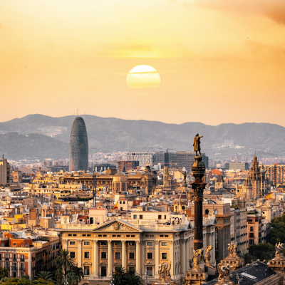 Barcelona cityscape with a prominent building and statue against a sunset sky.