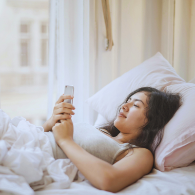 Woman lying in bed using a smartphone in a softly lit bedroom.